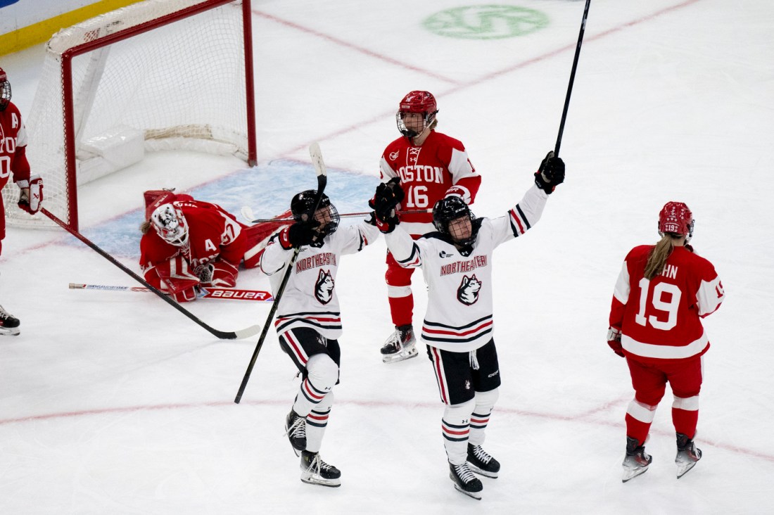 Northeastern womens hockey players celebrating a goal with their hands and sticks raised in the air.