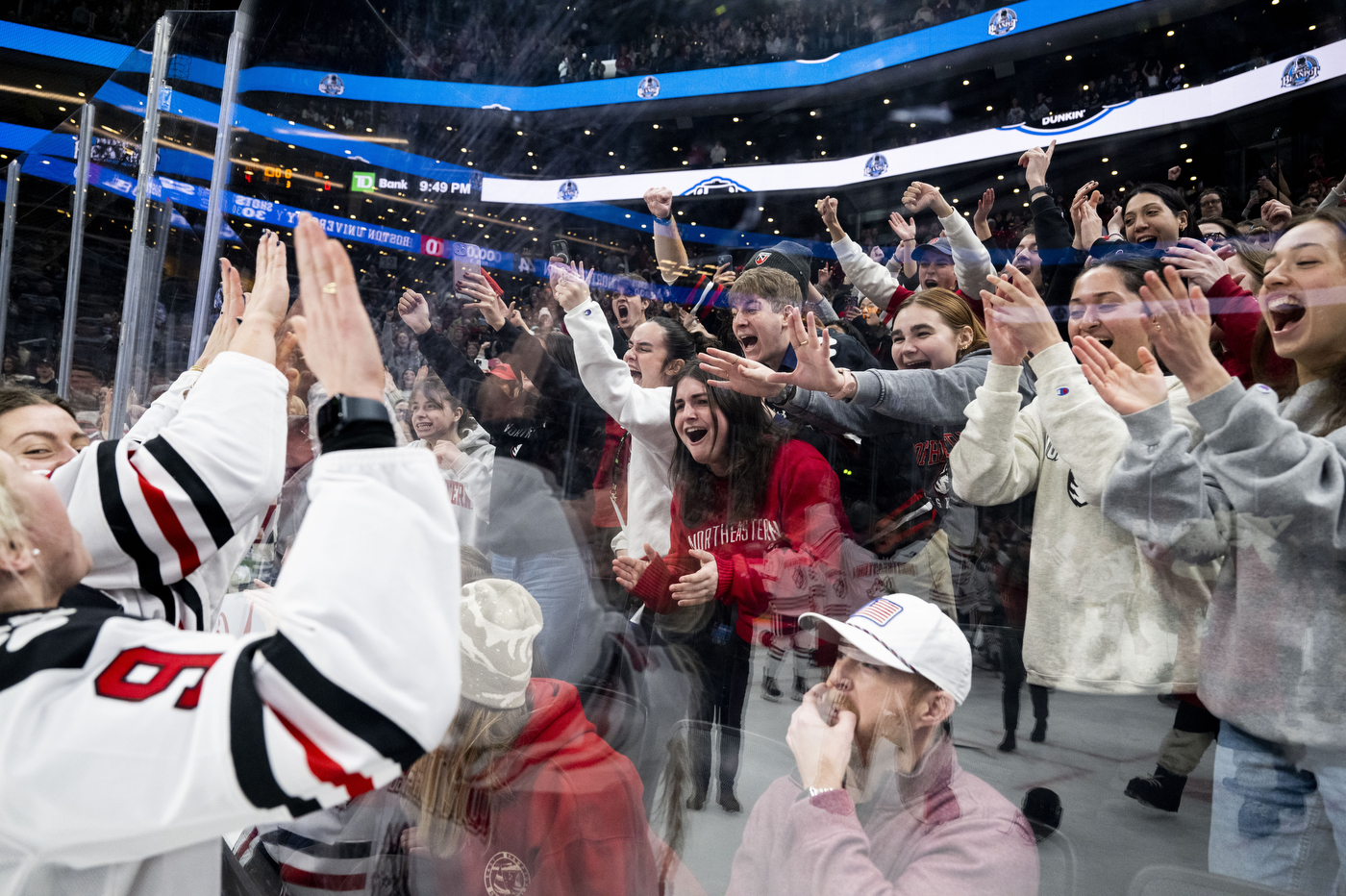 Northeastern hockey players celebrate with fans pressed against the glass at an arena. The crowd is cheering and reaching towards the team. 
