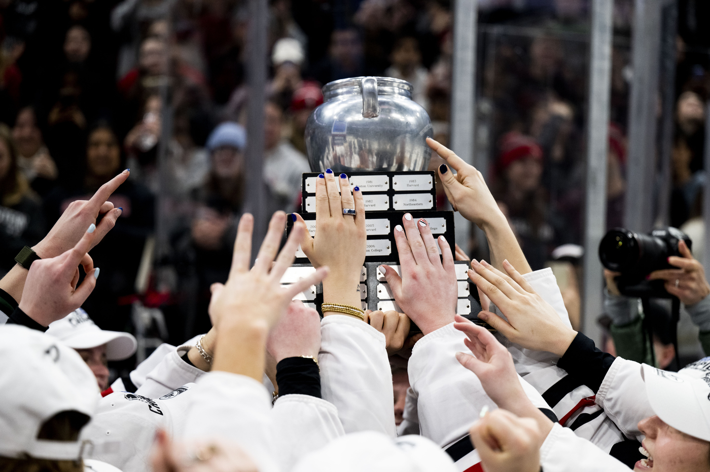 Northeastern hockey players hands reach up together to hoist the Beanpot championship trophy above their heads as the crowd celebrates in the background. 