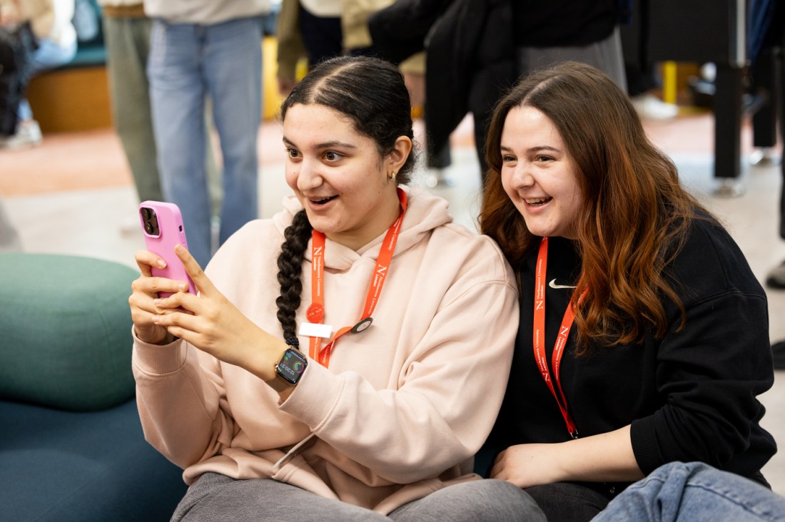 Two students sitting side by side, one holding up a pink iPhone.