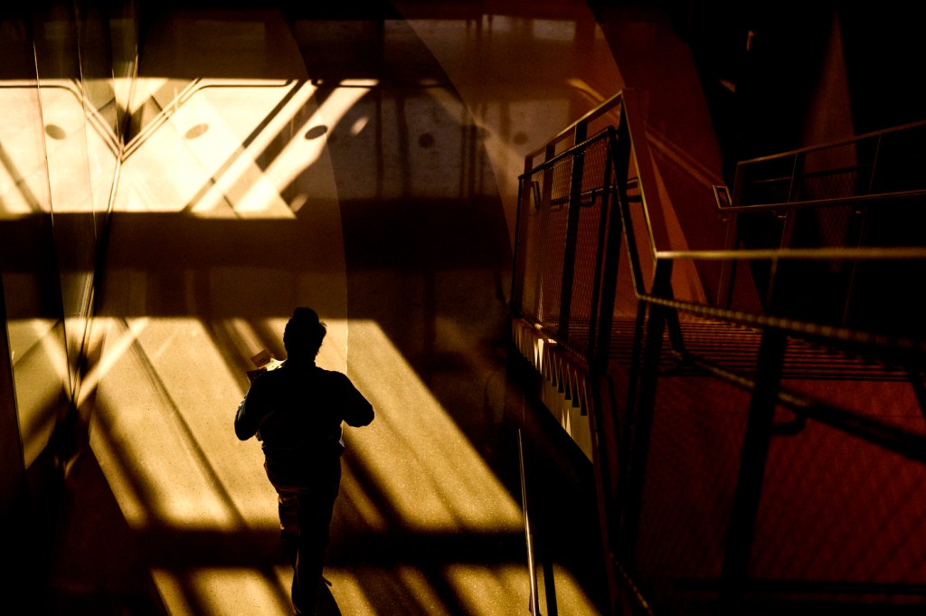 A figure walks through a sunlit space, casting long shadows against geometric patterns of light and structure.