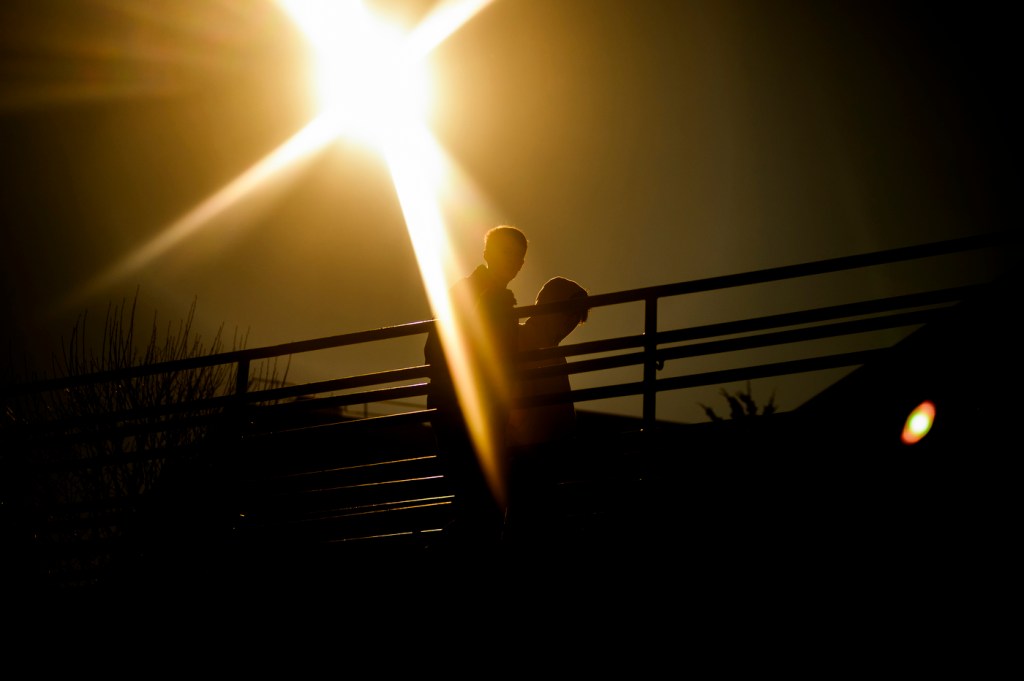 Silhouetted figures walk on a bridge against a bright, sunlit background.