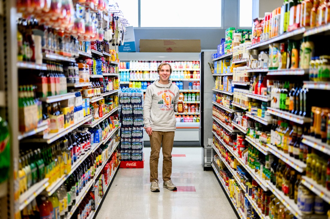 Jack Burns standing in a Wollaston's grocer in the condiment aisle wearing a sweatshirt that says 'Ketchup, Please' and holding a bottle of ketchup.