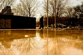 A figure walks past a reflective surface with bare trees and a somber monument in the background, evoking a quiet and contemplative mood.