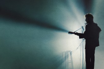 A screen capture from 'A Complete Unknown' of the silhouette of Timothee Chalamet as Bob Dylan playing guitar and singing on stage.