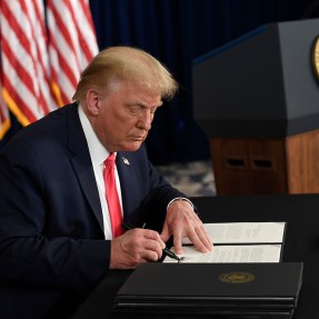 President Trump signing a piece of paper at a desk with US flags hanging behind him.