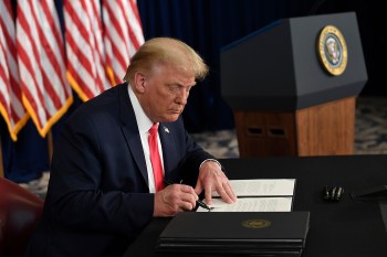 President Trump signing a piece of paper at a desk with US flags hanging behind him.