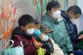 People sit indoors, wearing face masks, with a child holding a device in the foreground.