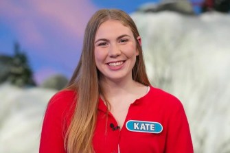 Portrait of Kate Stuntz on set at 'Wheel of Fortune' wearing a red shirt, a microphone, and a name tag.