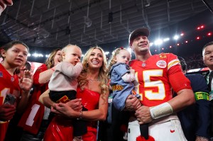 Patrick Mahomes wearing his Kansas City Chiefs Jersey, holding one of his kids. Next to him, his wife Brittany Mahones holds another one of their kids.