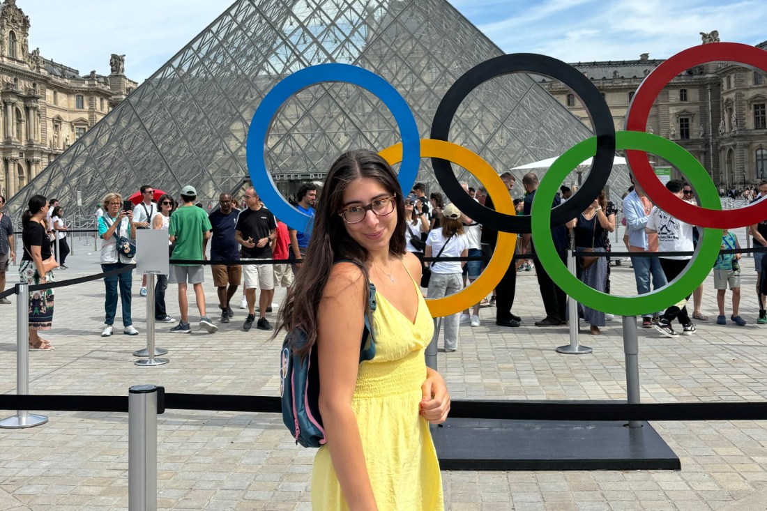 Mélanne Ghahraman wearing a yellow dress and a backback and glasses standing in front of the Olympic rings outside of the Louvre.