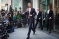 Prince Harry wearing a suit walking out of a building in front of photographers while waving a hand.