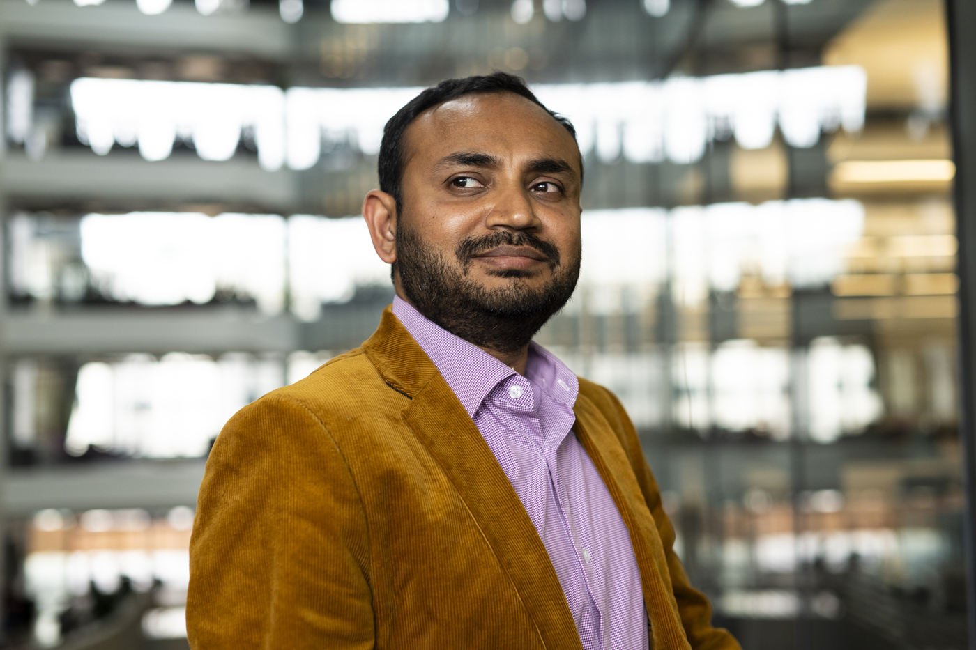 Portrait of Aanjhan Ranganathan wearing a pink button down shirt and a burnt orange velvet blazer, posing inside of a modern building. 