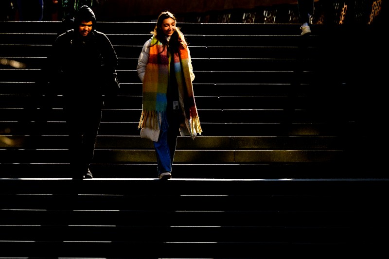 Two people walk down a set of stairs, illuminated by a stream of sunlight against a darker background.