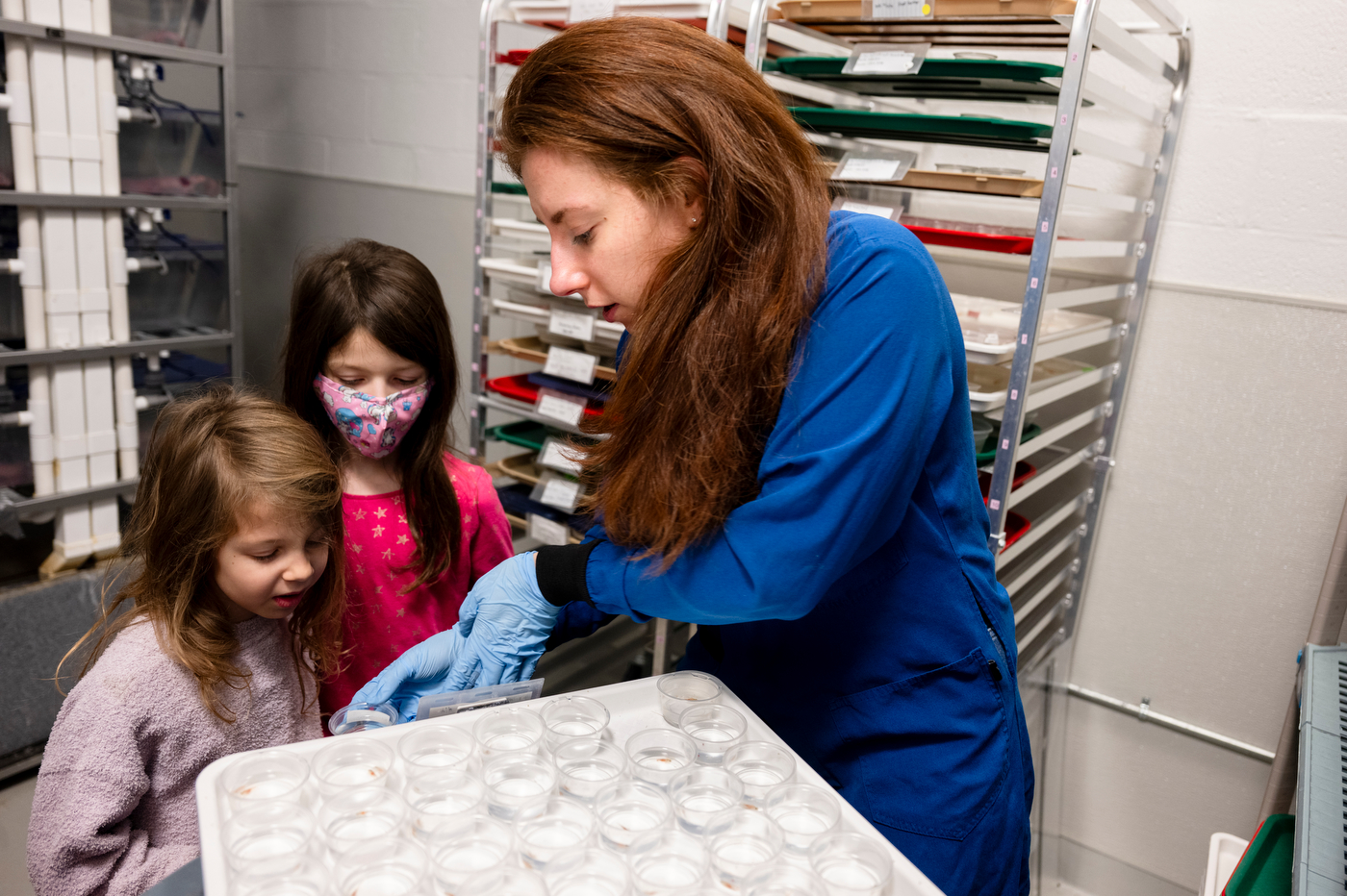 An individual in a lab coat engages with two children while examining small containers in a laboratory setting.