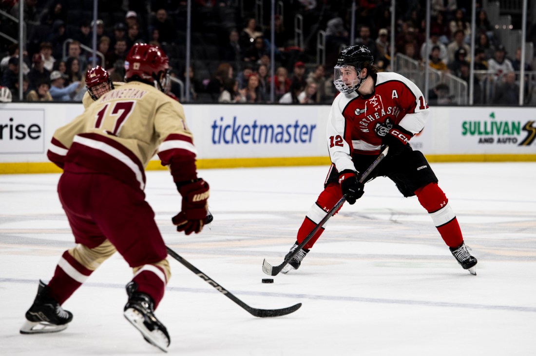 A Northeastern mens hockey player on the ice waiting to pass the puck. In front of him is a BC hockey player on defense.