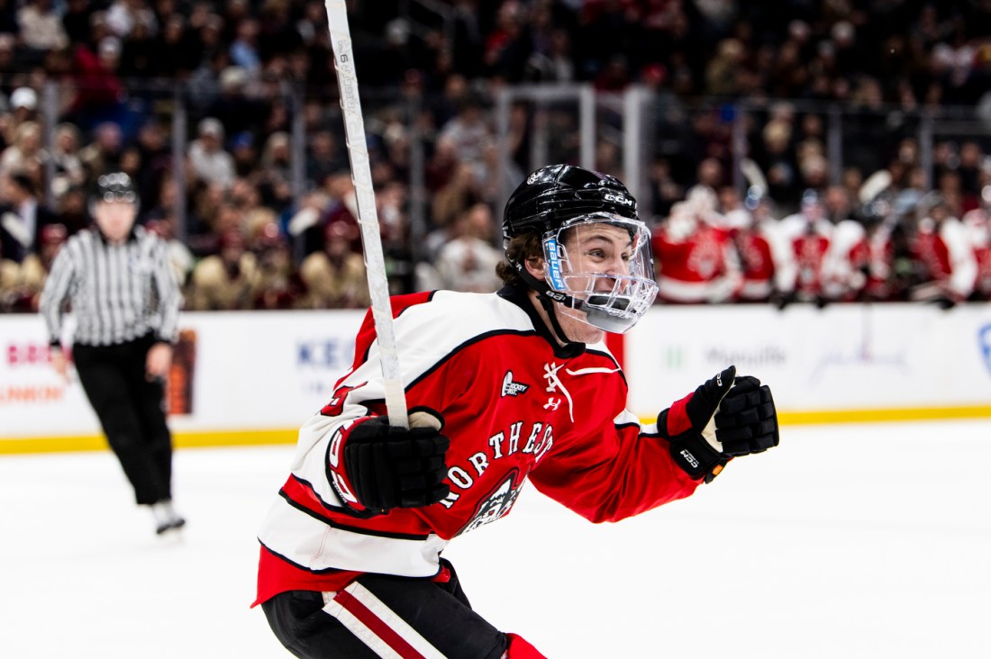 A Norhteastern mens hockey player cheering after scoring with his hockey stick up in the air. 