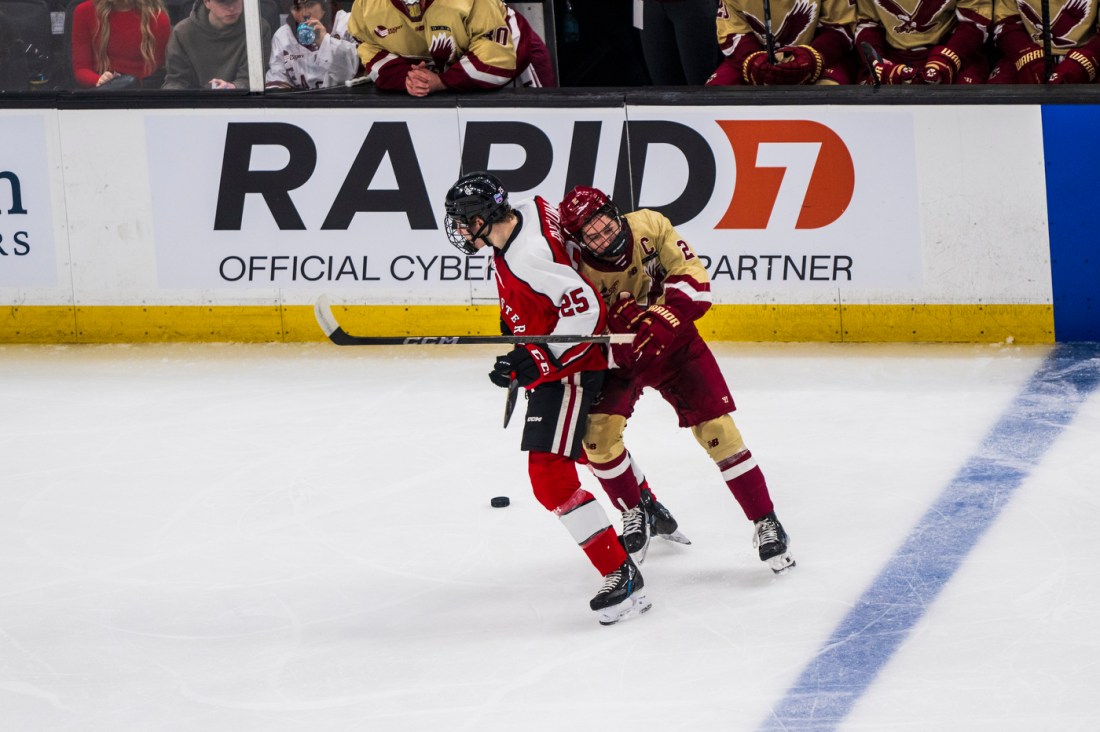 Two hockey players, one in a red and black jersey and the other in cream and maroon, closely battling near the boards.