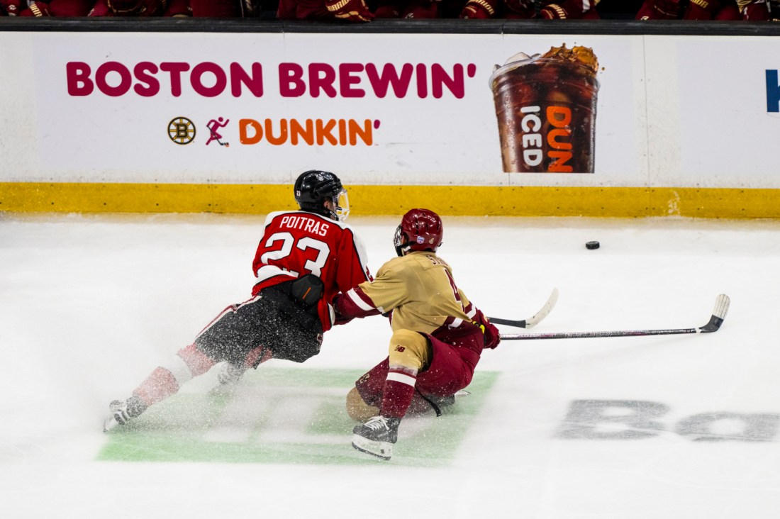 A player in a red and black jersey sliding on the ice while a cream and maroon player tries to intercept the puck.