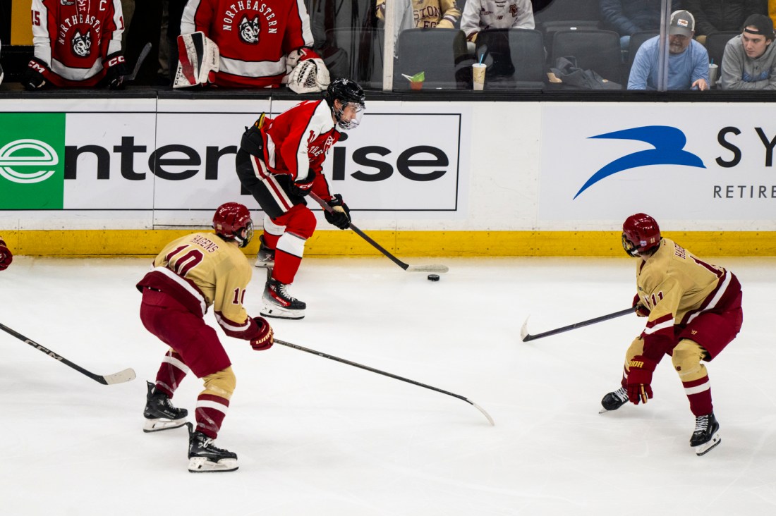 A player in a red and black jersey controlling the puck as two cream and maroon players skate towards them in defense.