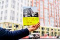 A hand holding up a printed image of a new building construction with a yellow sticky note on it saying "what we lost: our family homes, our views, our neighbors"