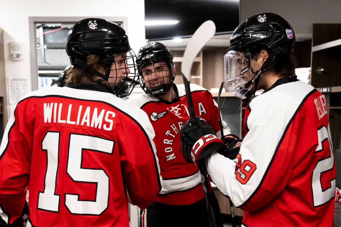 hree hockey players in red-and-white uniforms chat and laugh together in the locker room, holding their hockey sticks.