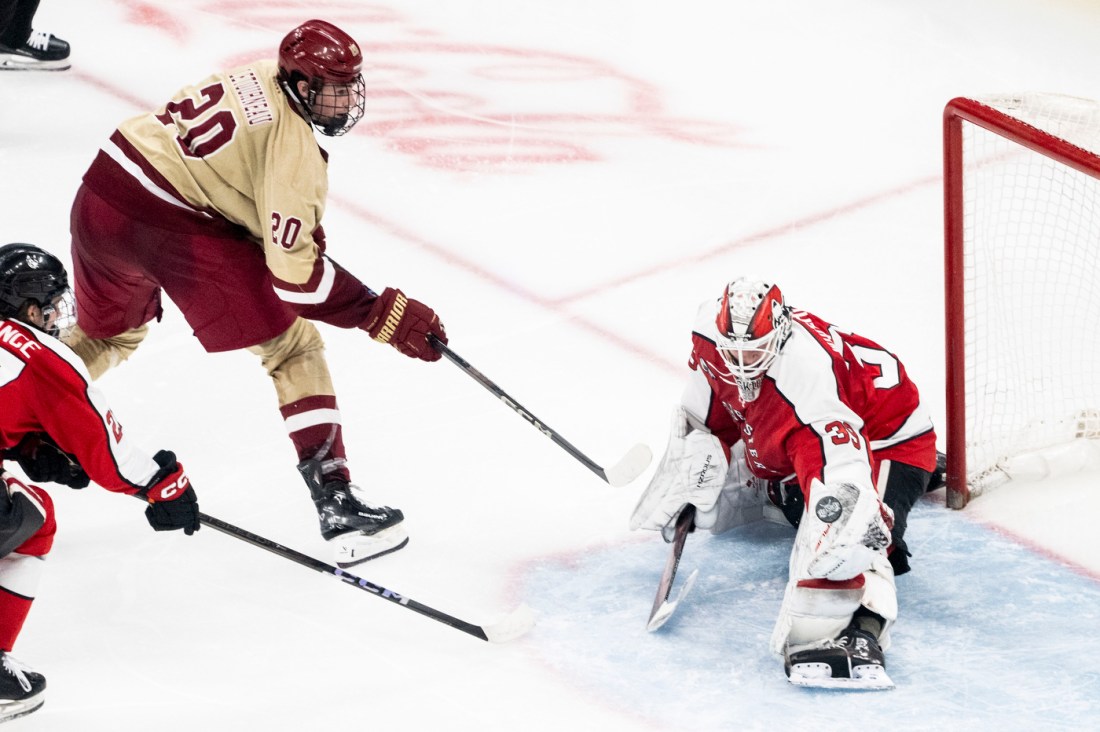 A player in a cream and maroon jersey attempting a shot on goal as a goalie in red and black crouches to block the puck.