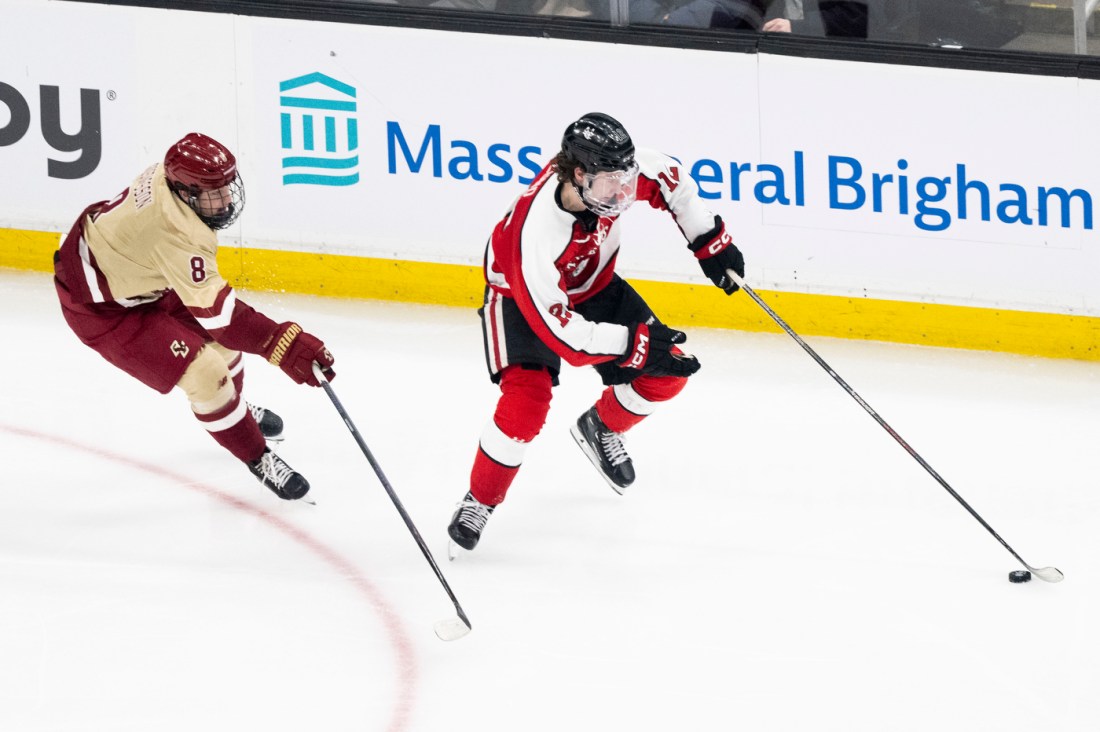 A player in a red and black jersey controlling the puck while being pursued by an opposing player in cream and maroon.