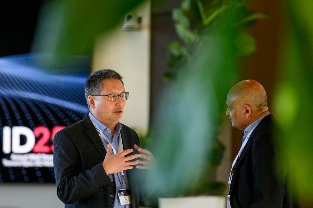 Two people wearing blue button downs and blazer jackets standing face to face. One is speaking while gesturing with his hands.