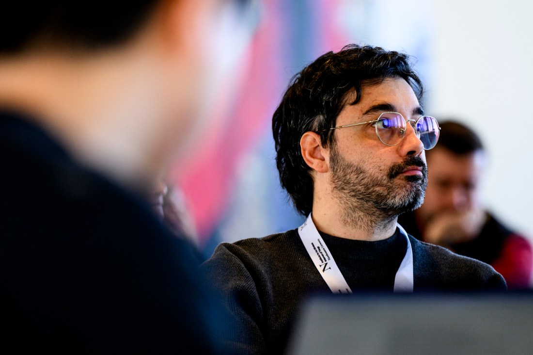 An audience member wearing a black shirt, glasses, and a white lanyard. 