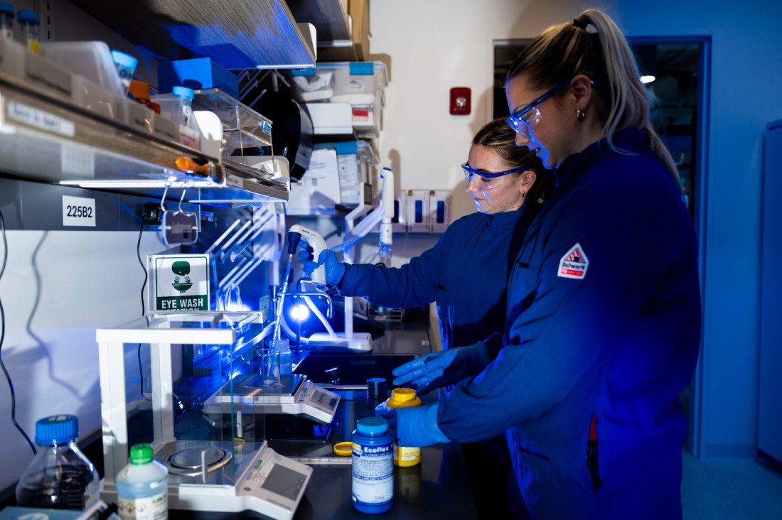 Two bioengineering students wearing lab coats and safety goggles standing in front of a counter with lab equipment on it. They are looking at a machine.