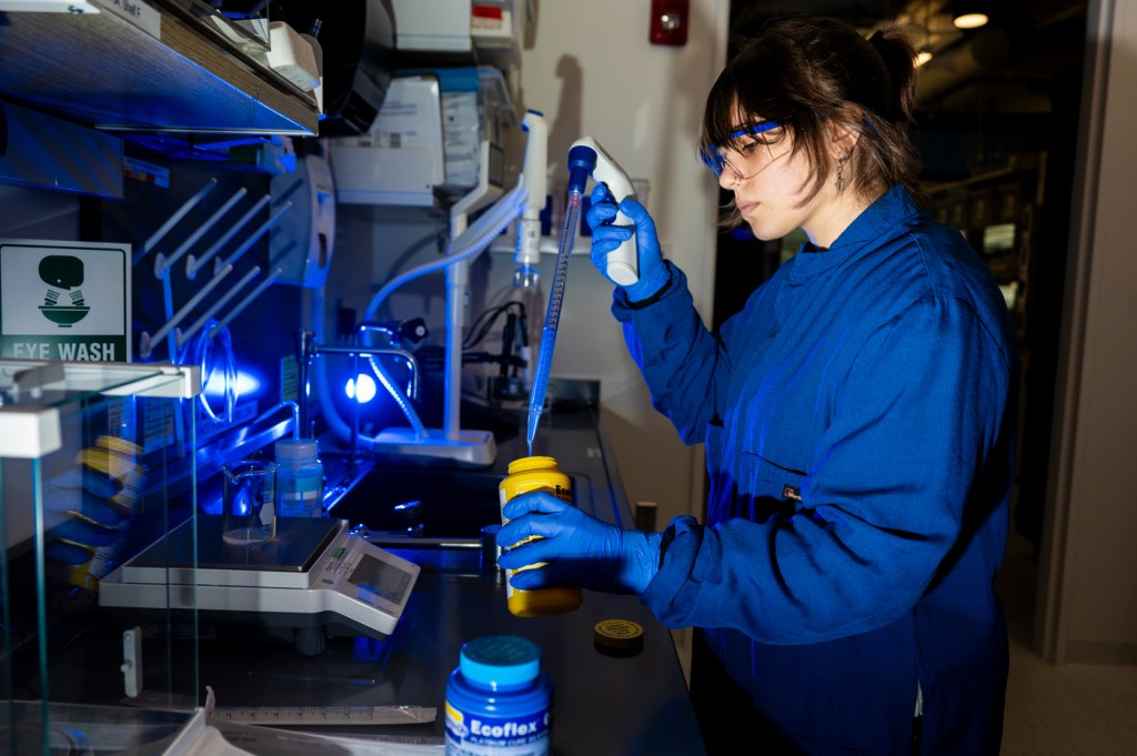 A person wearing safety goggles and a lab coat works with scientific equipment in a brightly lit laboratory.