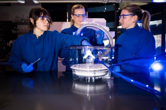 Three students wearing lab coats and safety glasses standing around a piece of equipment in a lab.