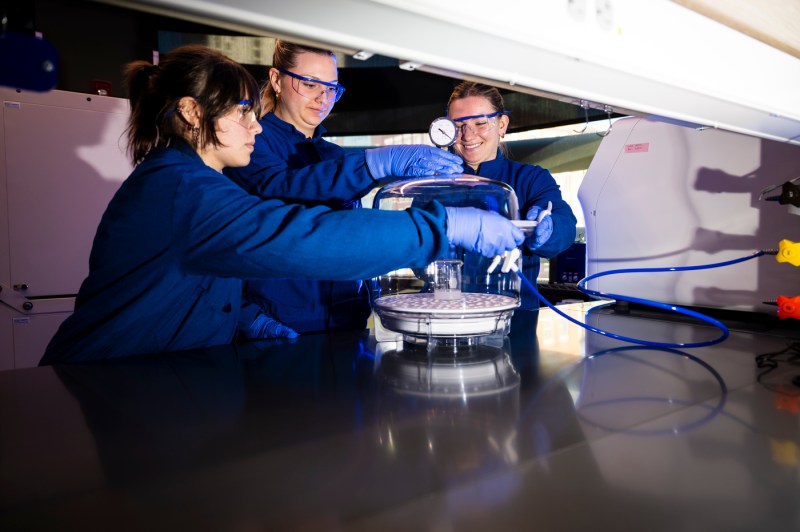 Three bioengineering students in a lab wearing lab coats and safety glasses standing around a piece of lab equipment.