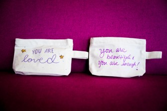 Two canvas bags laid out on a purple background. One says 'you are loved' on it and the other says 'you are beautiful! you are enough!' on it.