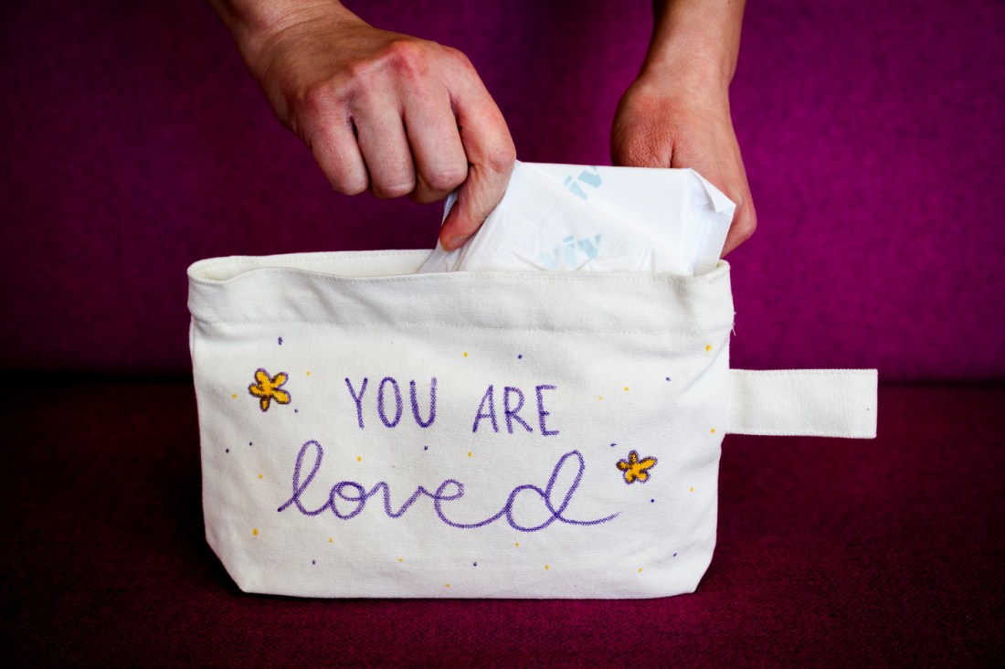 A set of hands pulling menstrual pads out of a canvas pouch that has 'you are loved' written on it in purple.