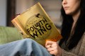 A person sitting on a green chair reading the book 'Fourth Wing' which has a gold cover and the title on it in black, all caps font.