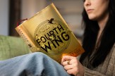 A person sitting on a green chair reading the book &#039;Fourth Wing&#039; which has a gold cover and the title on it in black, all caps font.