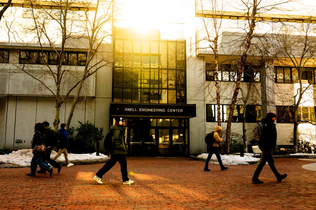 People walk along a brick path past a modern building, bathed in warm sunlight on a winter day.