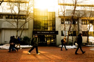 People walk along a brick path past a modern building, bathed in warm sunlight on a winter day.