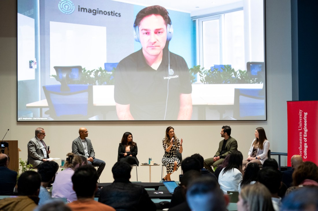 Six people sitting on stage underneath a projector screen. On the screen a person is sitting in a conference room wearing over the ear headphones.