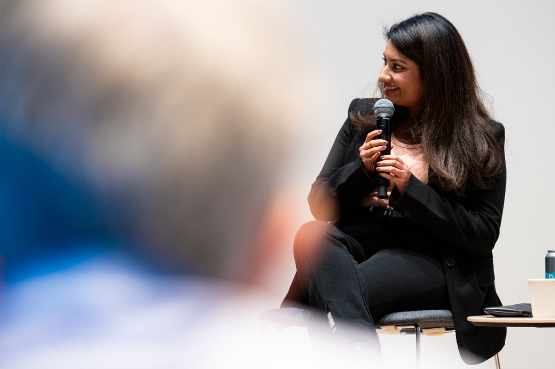 Shruti Kotian wearing black pants and a black blazer sitting in a chair holding a microphone.