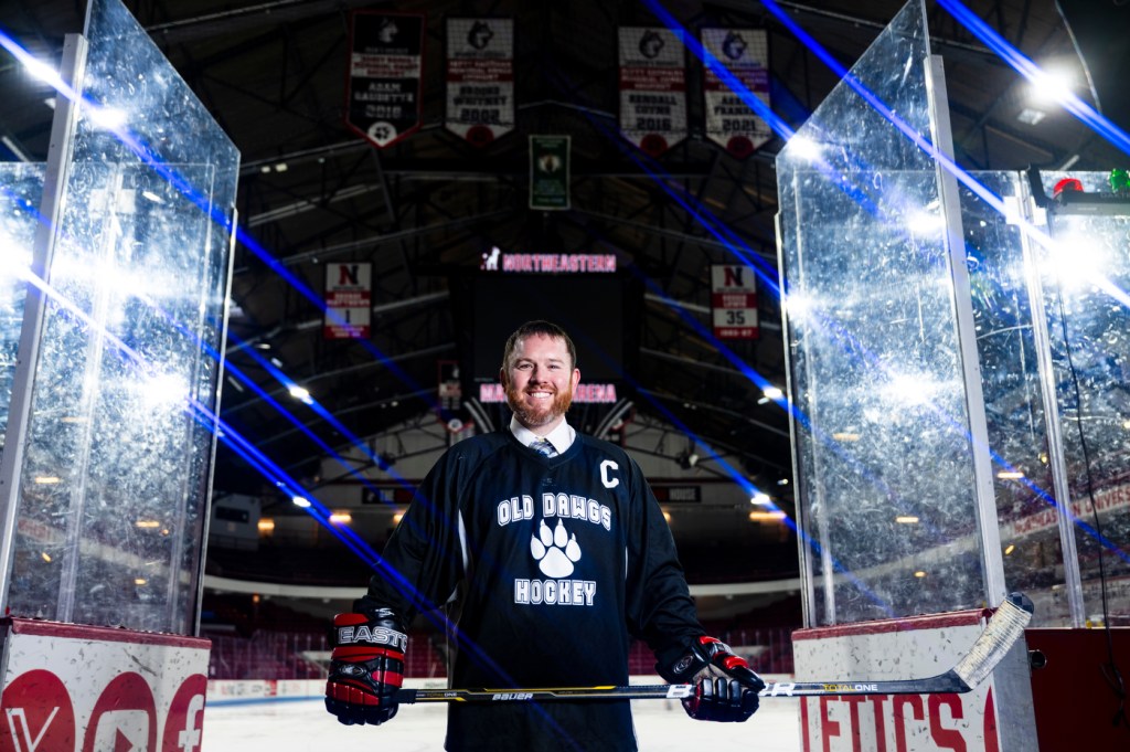 In a hockey jersey, Greg Smith stands on the ice at Matthews Arena holding his stick, with arena boards and banners visible in the background.