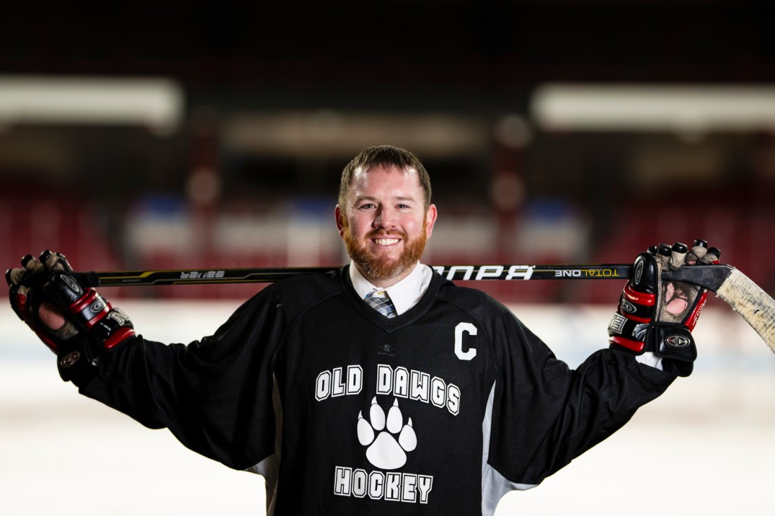 Greg Smith, wearing an "Old Dawgs Hockey" jersey and hockey gloves, holds his stick across his shoulders and smiles while standing on the ice at Matthews Arena.