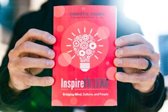 A person's hands holding Christie Chung's new book. It has a red cover with an illustration of a lightbulb and it has the title 'Inspire to Lead' on it.