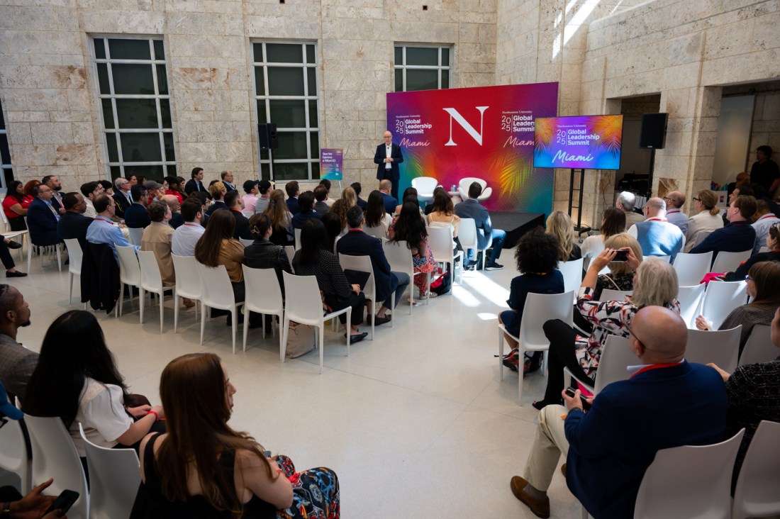 Rows of audience members sitting in white chairs in a room. Northeastern President Aoun is standing on stage at the front of the room in front of several screens that say '2025 Global Leadership Summit Miami'.