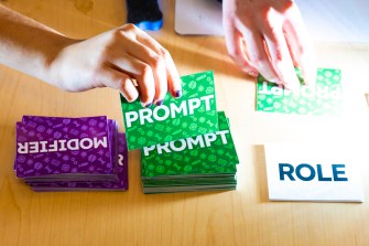 A person's hands picking up a card from a stack of cards labeled 'Prompt'. They're sitting next to a stack of cards labeled 'Modifier'.