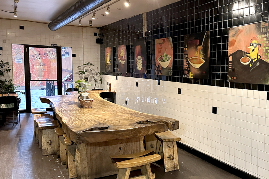 A large wooden table with stools around it inside of Ravi Soups. The wall behind the table is made of white tile on the bottom and black tile on the top, with paintings of different bowls of soup hanging on it. 