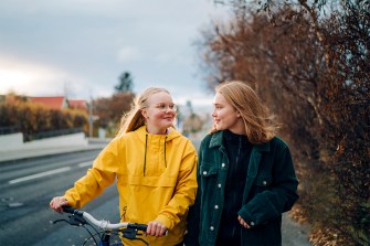 Two teenage girls looking at each other while walking down the street with a bike