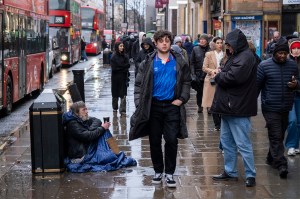 A man sitting on the pavement begging for money while shoppers walk past on a busy sidewalk.